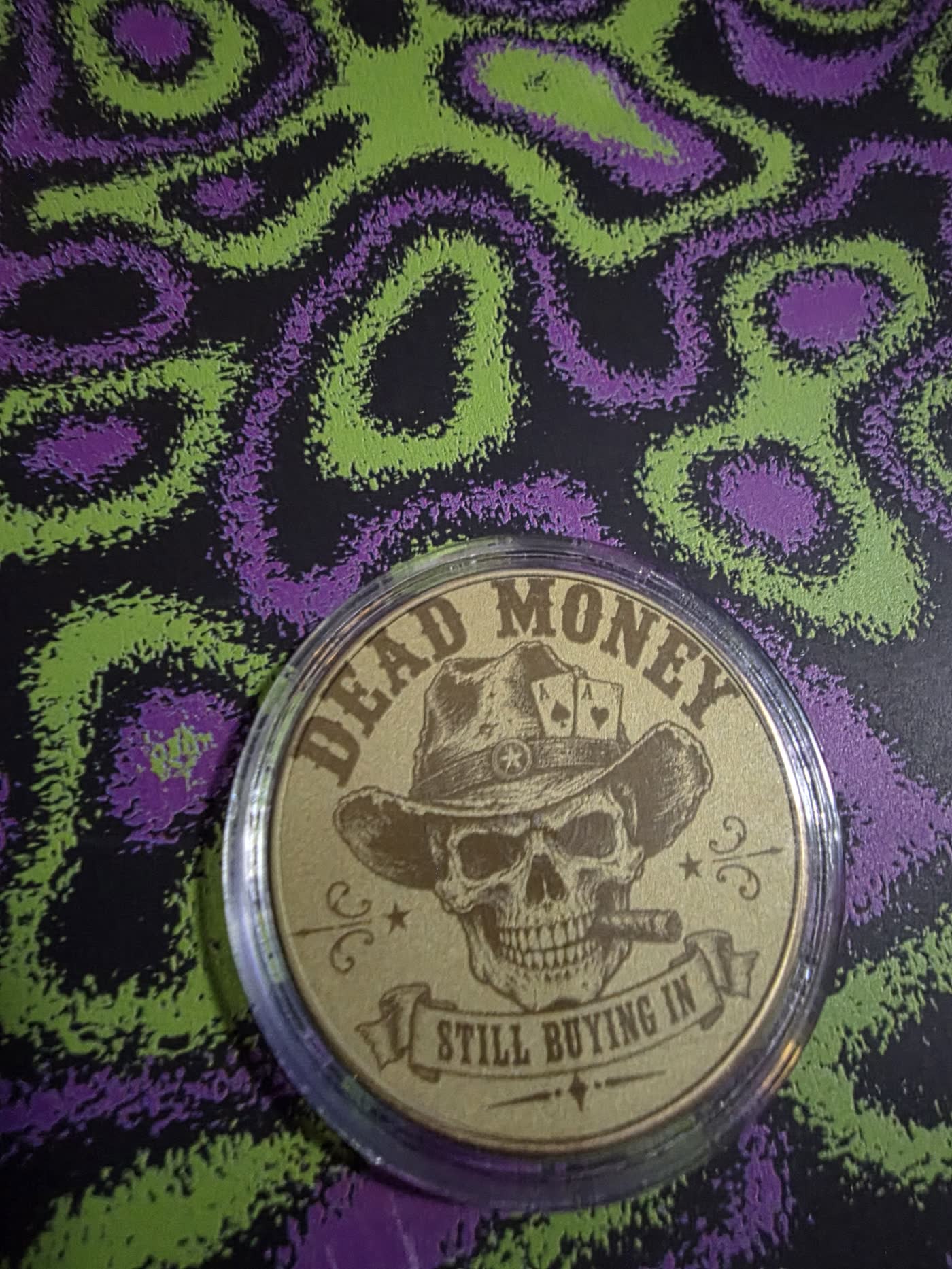 Gold metal poker card guard engraved with a cowboy skull smoking a cigar under the words Dead Money, Still Buying In.