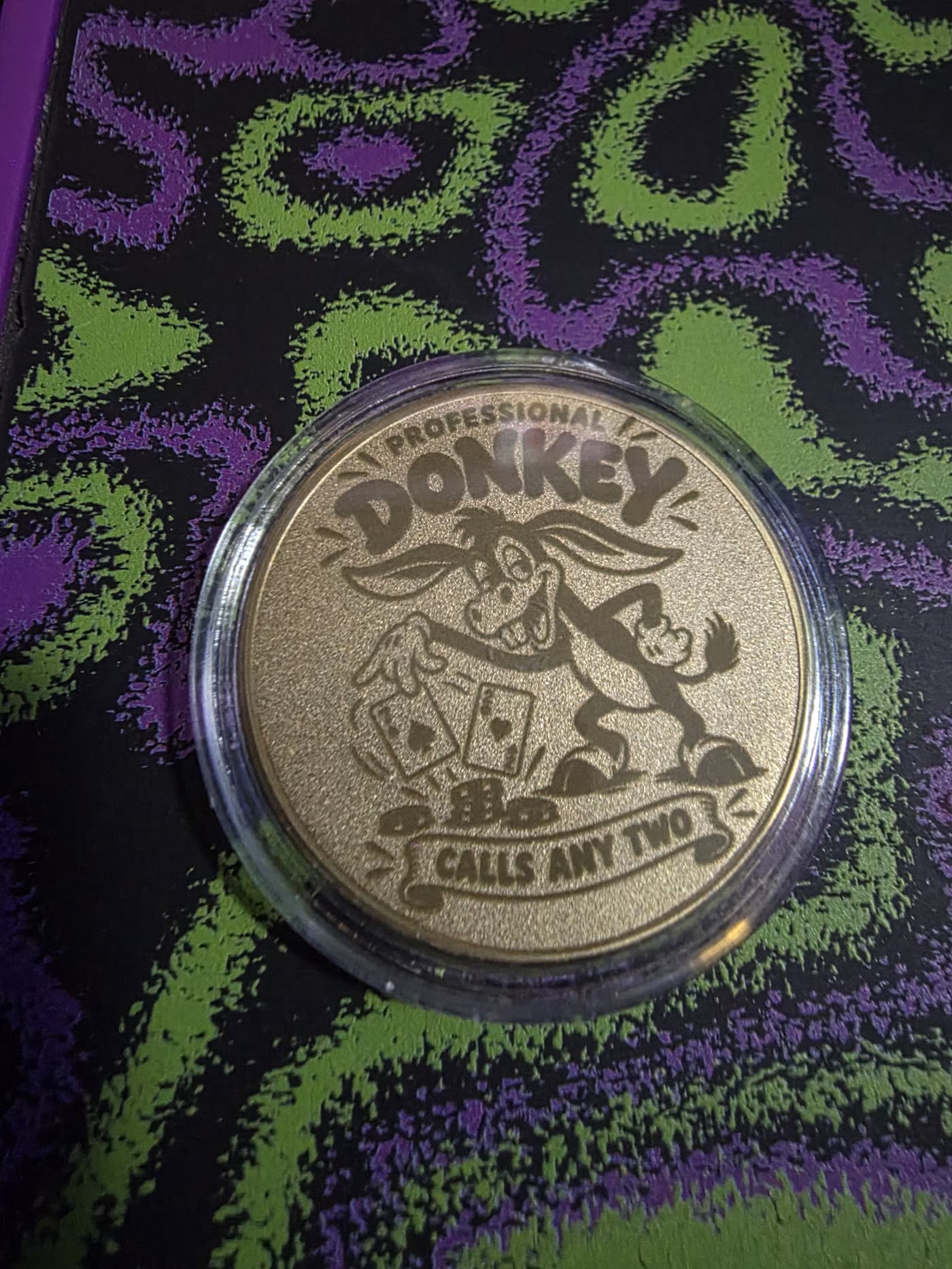 Gold metal poker card guard engraved with a laughing donkey holding two face cards and the line Calls Any Two.