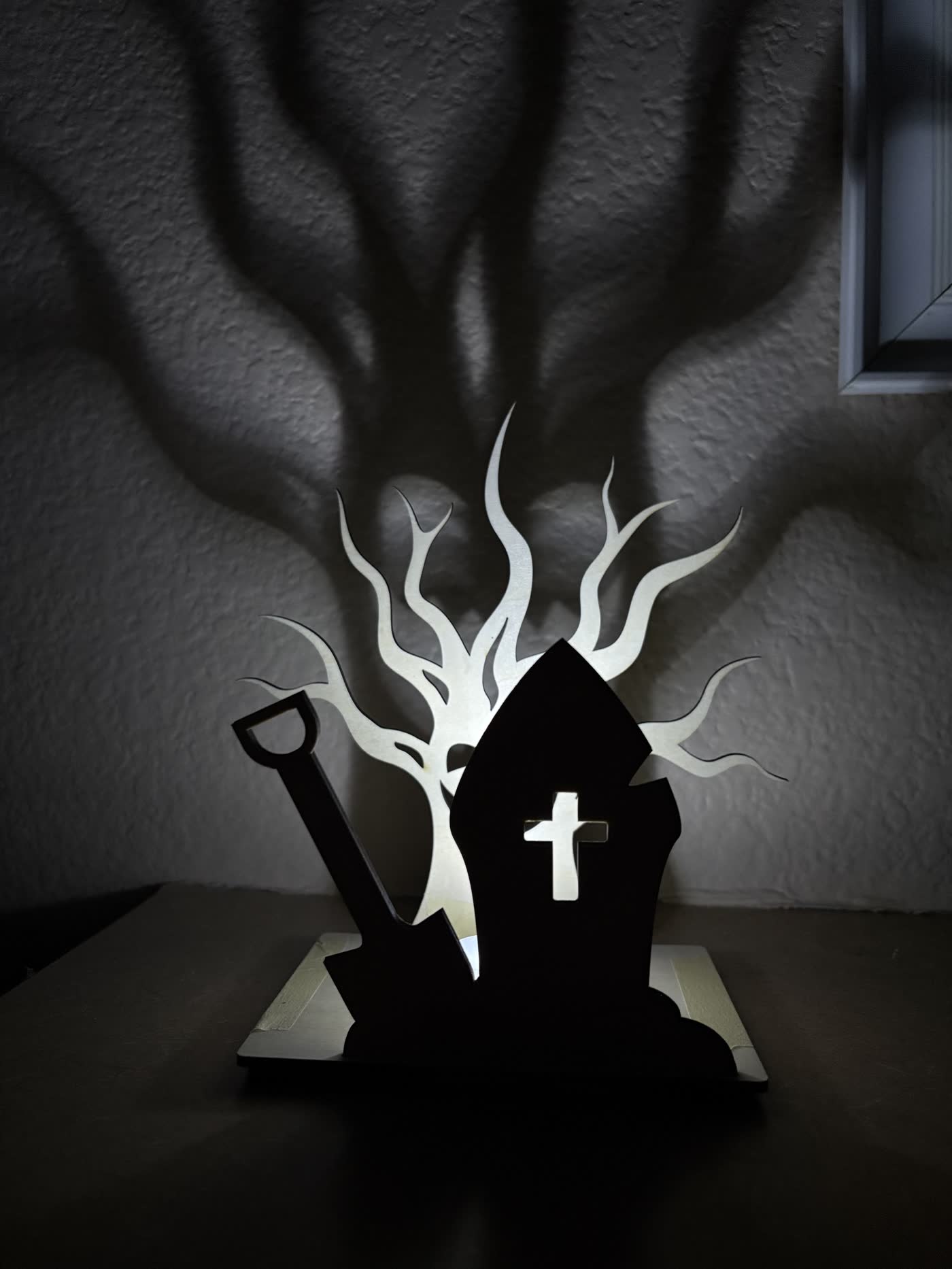 Wooden Halloween lantern with a shovel and headstone, casting a pale tree shadow on the wall.