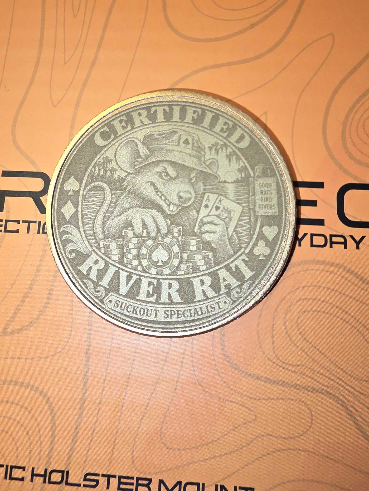 Brass metal poker card guard engraved with a grinning river rat in a fishing hat, holding pocket aces over a stack of chips.