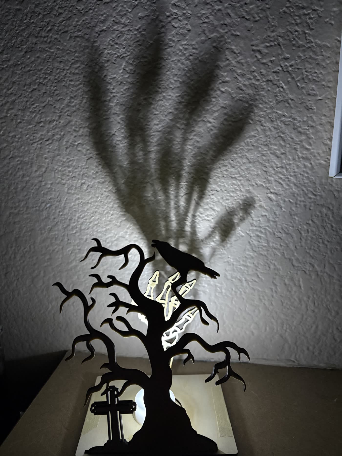 Wooden Halloween lantern of a leafless tree with a raven, casting a giant skeleton-hand shadow on the wall.